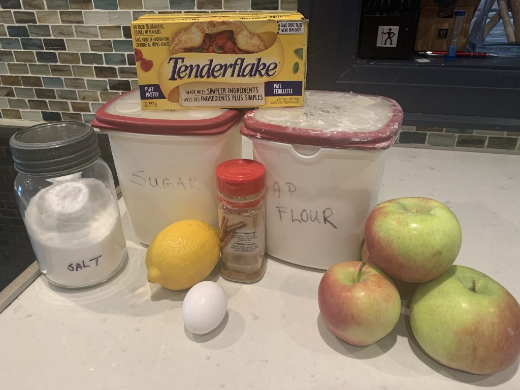 Picture of mason jar of salt; plastic tubs of sugar and flour; a lemon, an egg, a cinnamon jar, a pile of apples and tenderflake frozen puff pastry 