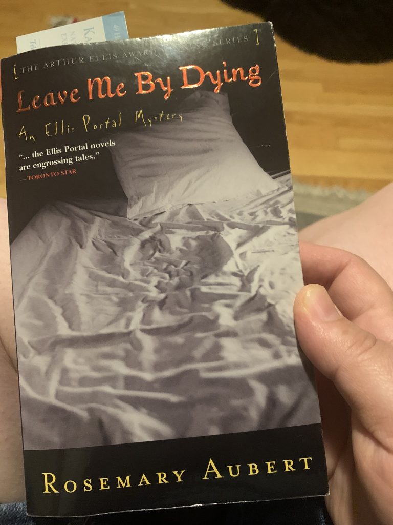 A picture of the cover of Leave Me by Dying which shows a rumbled white bed sheet and pillow. The book is being held in Lisa's right hand.