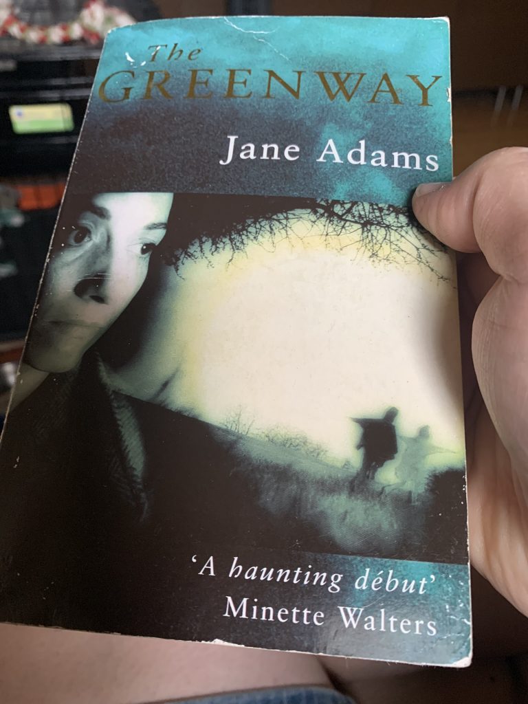 The picture shows the cover of The Greenway, a woman's face highlighted dramatically looks out over shadowy people. The book is held in Lisa's right hand.