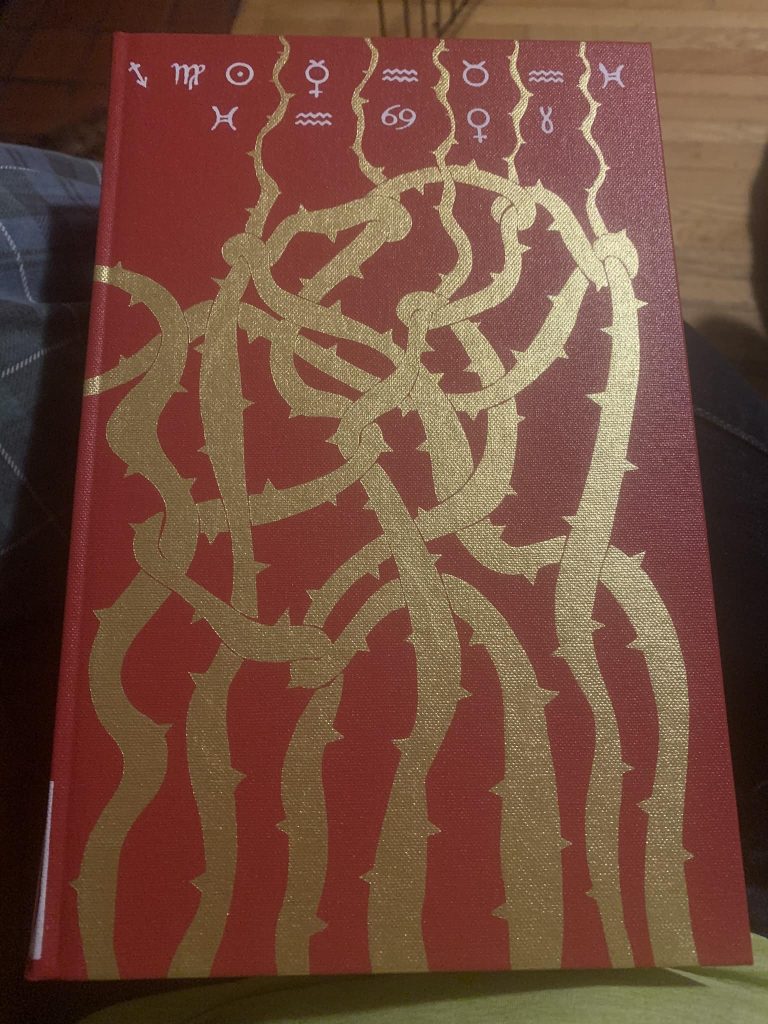A red hardcover book with thorn covered vines reaching towards the top from the bottom. Runes (astrological signs?) across the top.